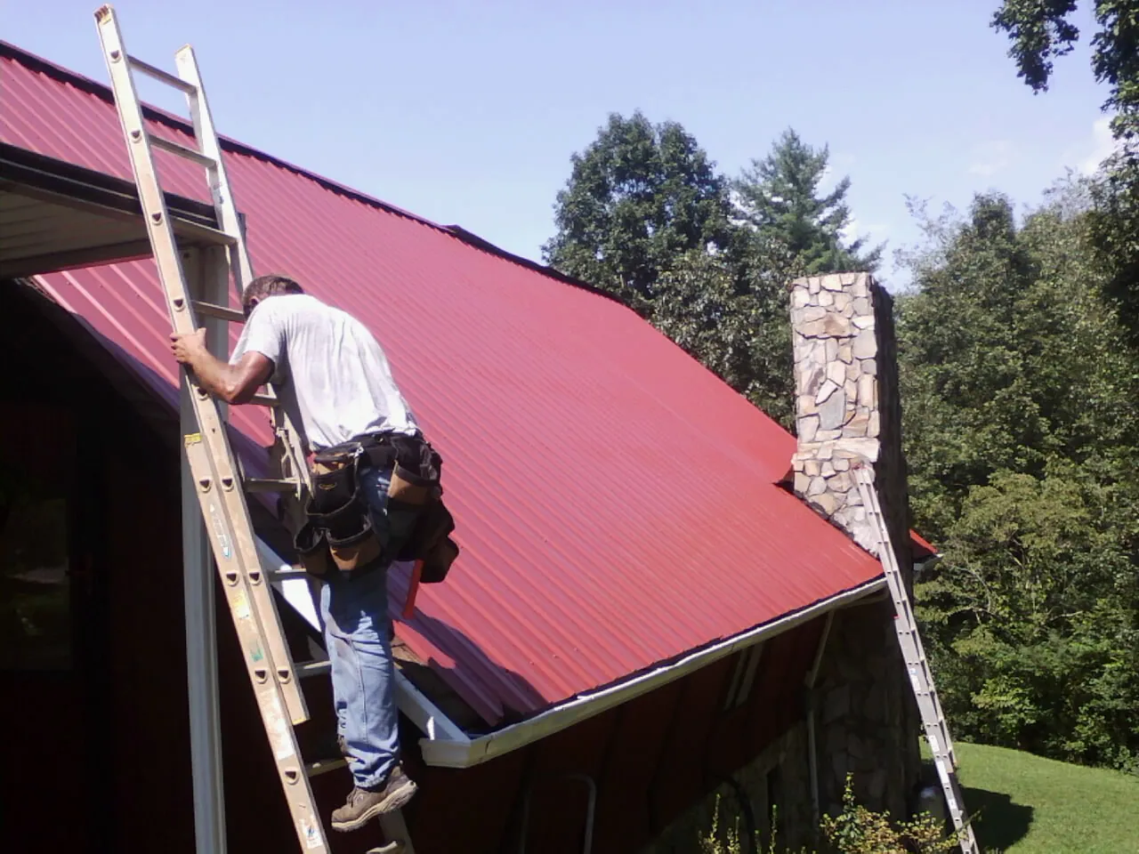 Roofer on a ladder beside a red metal roof and stone chimney — Soffit & Fascia Repair by Roofing Brothers in King And Queen Court House